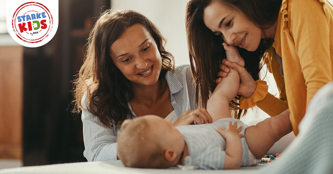 Zwei lächelnde Frauen spielen mit einem glücklichen Baby, das auf einem Bett liegt, in einem warm beleuchteten Zimmer.