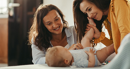 Zwei lächelnde Frauen spielen mit einem Baby, das auf einem Bett liegt.