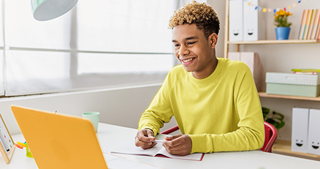 Ein fröhlicher Teenager mit lockigem Haar und einem gelben Pullover sitzt an einem Schreibtisch, lächelt und schreibt etwas, während er auf einen Laptop-Bildschirm schaut.